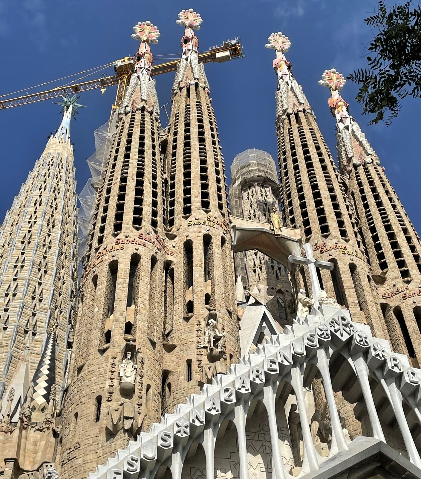 Fotografia wykonana z żabiej perspektywy przedstawia cztery główne i jedną boczną wieżę kościoła Sagrada Familia (świętej rodziny) w Barcelonie. To nietypowe rozwiązanie konstrukcyjne, łączące organiczną fakturę powierzchni budowli, pełną ażurów i różnorodnych struktur z majestatyczną wysokością. Na pierwszym planie wybija się fragment trójkątnego portalu z ozdobnym, koronkowym gzymsem, który wspiera rząd cienkich kolumienek. Za wieżami wystaje ramię dźwigu, które przypomina o niezakończonej budowie świątyni.