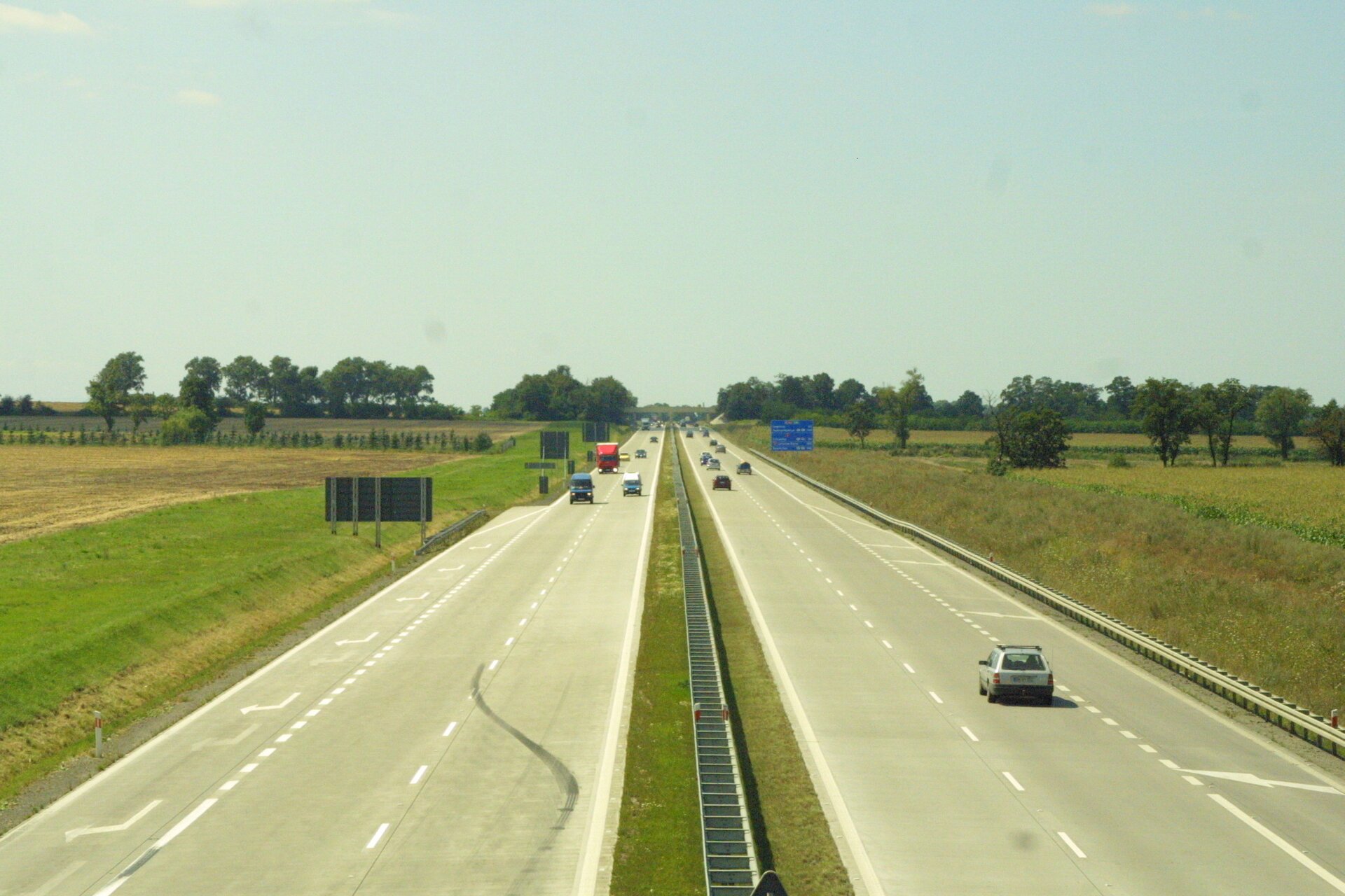 Na zdjęciu autostrada, dwa pasy i pobocze w każdym kierunku. Po obu stronach pola uprawne.