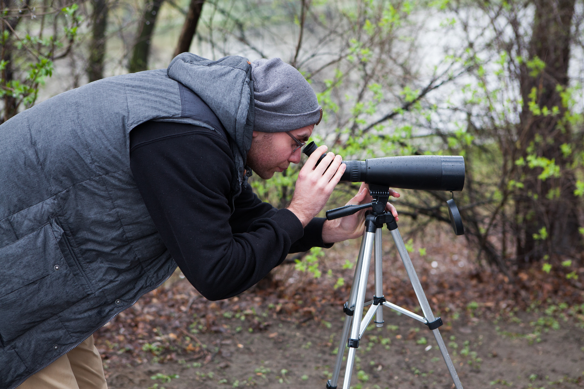 Fotografia ukazuje człowieka patrzącego przez lunetę na statywie. Mężczyzna ubrany jest w szarą czapkę, szarą kamizelkę i czarną bluzę oraz ma założone okulary. W tle znajdują się drzewa i krzewy.