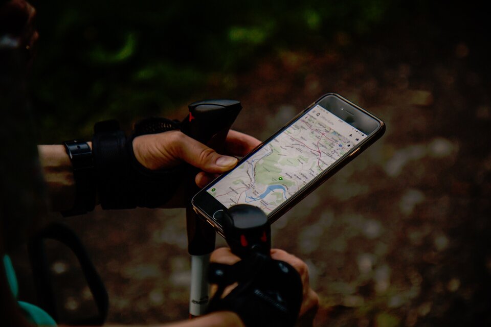 Fotografia przedstawiająca kierownicę rowerową, pośrodku której za pomocą uchwytu zamontowany jest smartfon. Na jego ekranie wyświetla się ekran nawigacji GPS. Przedstawiona jest na nim siatka ulic.