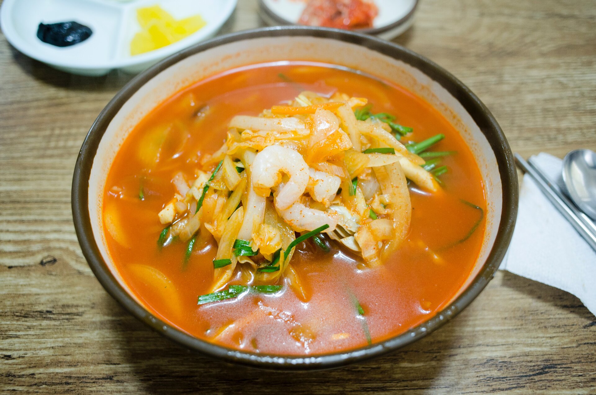 Zdjęcie przedstawia miskę z zupą. W czerwonym wywarze znajdują się krewetki oraz warzywa. Obok leżą sztućce.