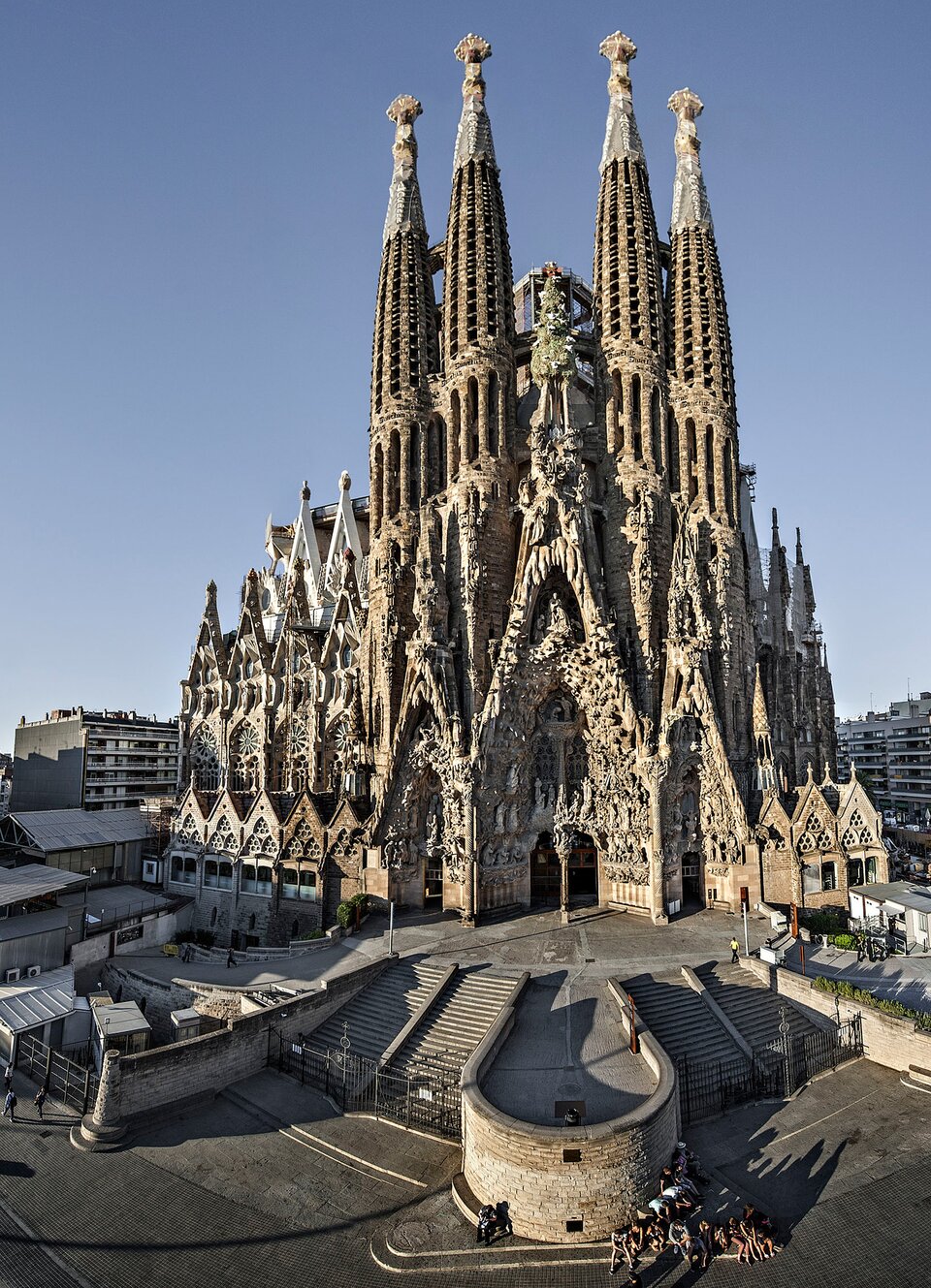 Ilustracja przedstawia kościół Sagrada Familia z Barcelony, zaprojektowany przez Antoniego Gaudíego. Ukazuje budowlę o charakterystycznych, strzelistych wieżach, których powierzchnie są gęsto pokryte rzeźbiarskimi detalami. Fasada widoczna na zdjęciu sprawia wrażenie niemal wyrastającej z kamienia — jej forma jest pełna nieregularnych, organicznych kształtów, typowych dla stylu Gaudiego. W dolnej części fotografii widać wejście prowadzące do świątyni, otoczone schodami i balustradą o miękkiej, falującej linii. Przed budowlą stoją ludzie, co podkreśla monumentalną skalę obiektu. W tle dostrzec można zabudowę miejską Barcelony, która kontrastuje z niezwykłą, fantazyjną formą bazyliki.