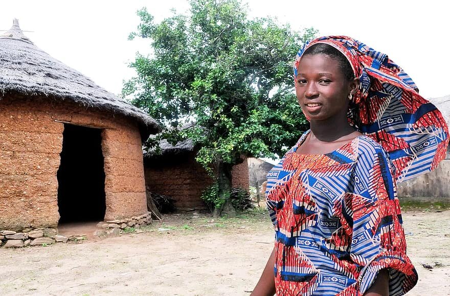 Zdjęcie przedstawia młodą, czarnoskórą kobietę stojącą nieopodal domu. Kobieta ma kolorową chustkę na głowie oraz ubrana jest w sukienkę z tego samego materiału co chustka. Wygląda bardzo schludnie. Dom jest niski, okrągły, ma kamienną podstawę, dach w kształcie stożka. Z przodu domu jest wycięty otwór w kształcie drzwi. Obok domu rośnie drzewo.   