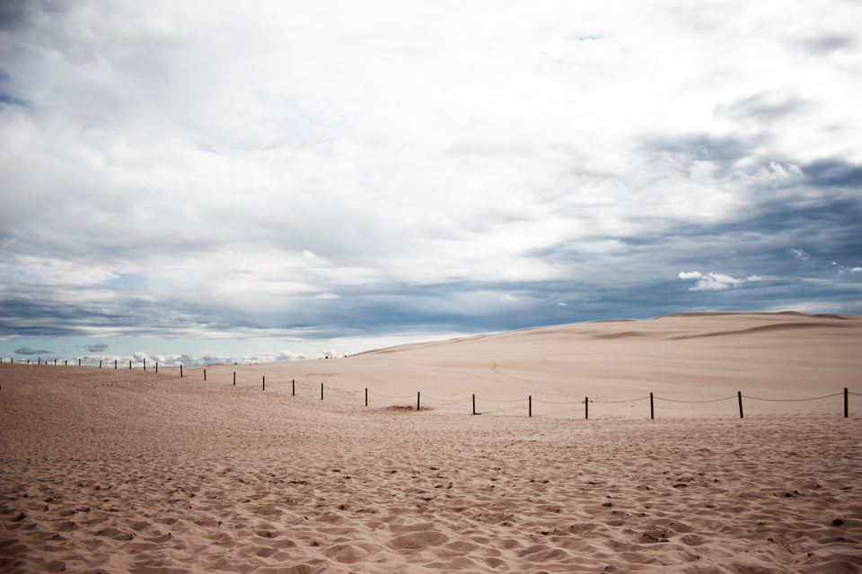 Fotografia przedstawia piaszczyste wydmy pod zachmurzonym niebem. To ruchome wydmy w Słowińskim Parku Narodowym. Przez wydmy biegnie ogrodzenie z palików.
