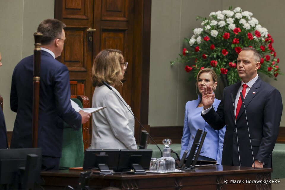 Ilustracja przedstawia uroczystą scenę w sali sejmowej.
Po prawej stronie stoi mężczyzna w garniturze z czerwoną krawatą i małą przypinką w barwach Polski w klapie marynarki. Unosi prawą rękę, jakby składał przysięgę lub ślubowanie. Obok niego, nieco w tle, stoi kobieta w jasnoniebieskim stroju, spokojnie patrząca w stronę przysięgającego.
Po lewej stronie kadru znajdują się dwie inne osoby – mężczyzna w garniturze trzymający dokumenty oraz kobieta w białej marynarce, którzy prowadzą ceremonię.
W tle, na ścianie, widać duży bukiet biało‑czerwonych kwiatów – symboliczne kolory flagi Polski. Scena odbywa się w poważnej, urzędowej atmosferze.