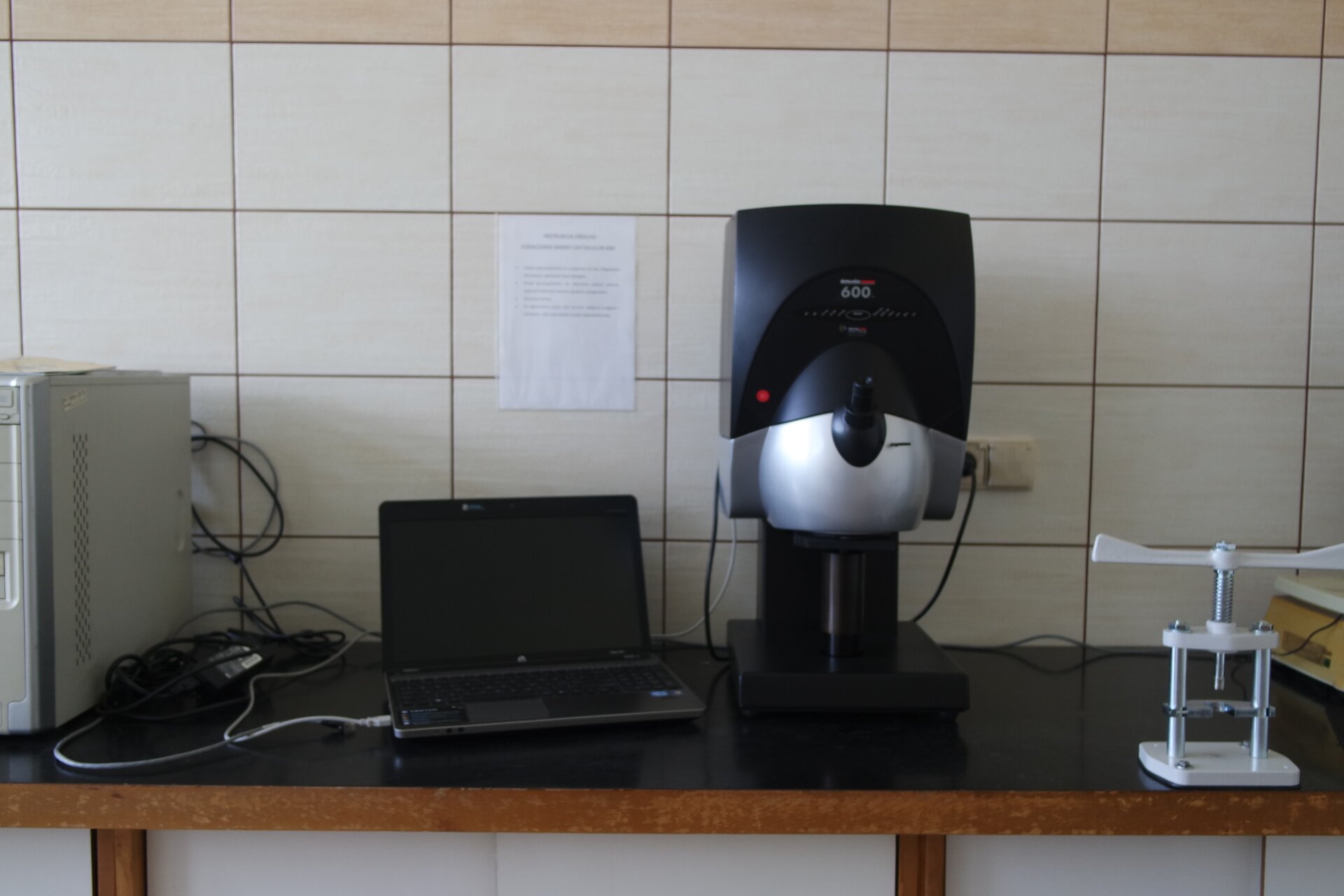 Zdjęcie ukazuje spektrofotometr do pomiaru barwy. Urządzenie składa się z szaro‑stalowej części głównej, która podłączona jest do komputera usytuowanego po lewej stronie grafiki. Po stronie prawej widoczny jest biały przyrząd przypominający wiatraczek.
