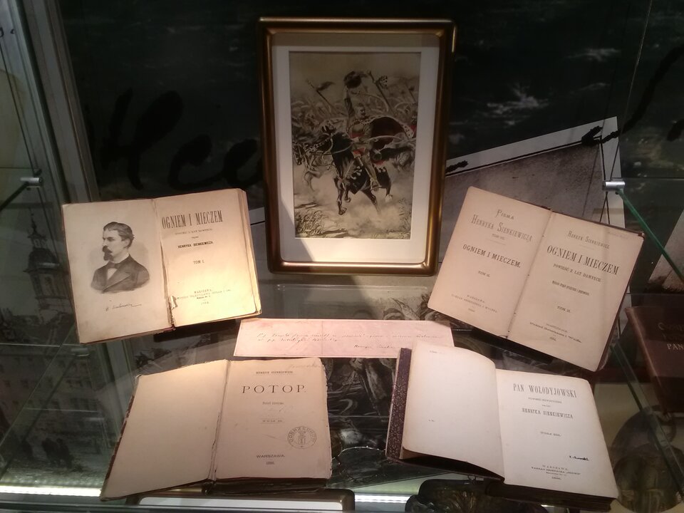 Zdjęcie przedstawia gablotę, w której znajduje się wystawa książek Henryka Sienkiewicza: "Ogniem i mieczem", "Potop" oraz "Pan Wołodyjowski". W gablocie stoi również oprawione w ramkę zdjęcie jeźdźcy na koniu.