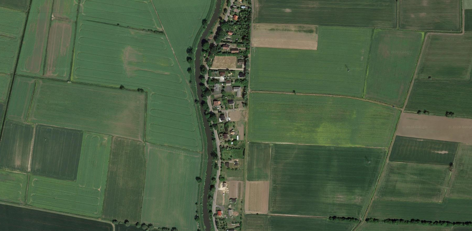 Zdjęcie przedstawia widok z góry na płaski teren z licznymi polami. Na środku zdjęcia jest wąski zabudowany pas, po jego lewej stronie biegnie droga, a dalej jest rzeka.   