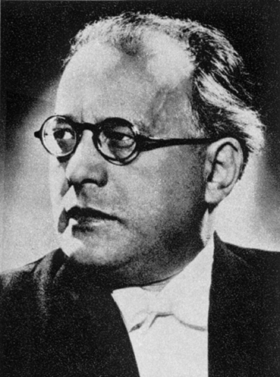 Ilustracja przedstawia portret Grzegorza Fitelberga - polskiego dyrygenta, kompozytora i skrzypka. Mężczyzna w średnim wieku, ma duże zakola i nosi okulary z okrągłymi oprawkami. Ubrany jest w białą koszulę i muszkę oraz ciemną marynarkę. Zdjęcie jest czarno‑białe.