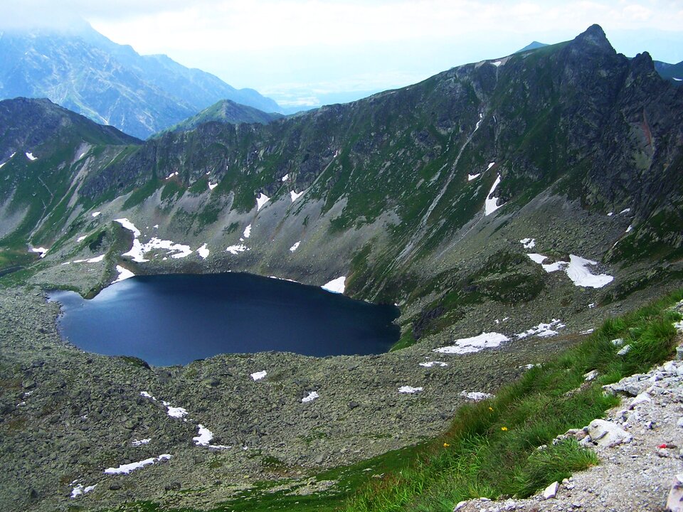 Zdjęcie ukazuje jezioro z ciemną wodą, znajdujące się u stóp wysokiej góry o ostro schodzących w kierunku jeziora zboczach. Tu i ówdzie leży plama śniegu.  