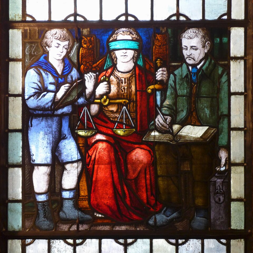 Ilustracja przedstawia witraż ukazujący trzy postaci. Po lewej stronie stoi chłopiec. Ma gęste, falujące włosy okalające okrągłą twarz, lekko przymknięte oczy, mały nos i pełne usta. Pochyla lekko głowę. Jest ubrany w koszulę z szerokim, marynarskim kołnierzem i krótkie spodnie sięgające kolan. Na nogach ma długie skarpety i wiązane z przodu buty. W lewej ręce trzyma tablicę do notowania. W prawej dłoni trzyma rysik. Notuje. Pośrodku na ozdobnym tronie siedzi dojrzała kobieta. Ma falujące włosy, owalną twarz, opaskę zakrywającą oczy, mały nos i pełne usta. Ubrana jest w ozdobną suknię z napisem Justitia. Na nią narzucono obszerną pelerynę. Spod peleryny wystaje fragment bosej prawej stopy. W prawej dłoni trzyma wagę. Lewą rękę opiera o rękojeść miecza. Po prawej stronie siedzi dojrzały mężczyzna. Ma falujące włosy, owalną twarz, lekko zmarszczone brwi, głęboko osadzone oczy, gęste wąsy. Lekko pochyla głowę. Ubrany jest w koszulę, kamizelkę i surdut. Na nogach ma spodnie i wsuwane pantofle. Na kolanach trzyma otwartą grubą księgę. Notuje w niej. W lewej dłoni trzyma monetę, którą wrzuca do otworu w stojącej pod jego nogami skarbonki.