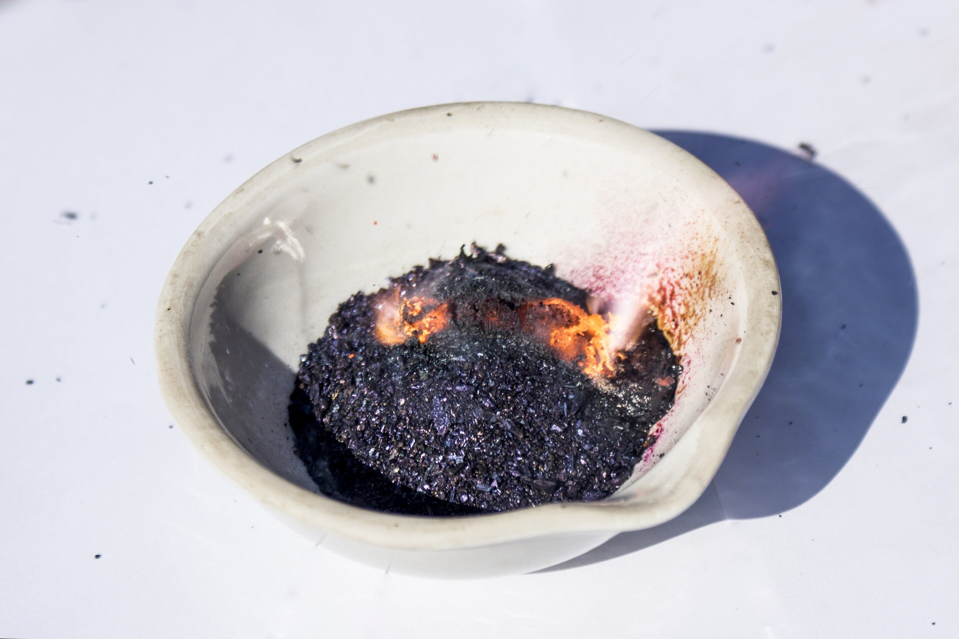 Zdjęcie przedstawia czarno‑fioletowy krystaliczny, lekko połyskujący manganian(<math aria‑label="siedem">VII) potasu znajdujący się w białej porcelanowej parowniczce wraz z glicerolem. Zawartość parowniczki pali się pomarańczowym płomieniem.