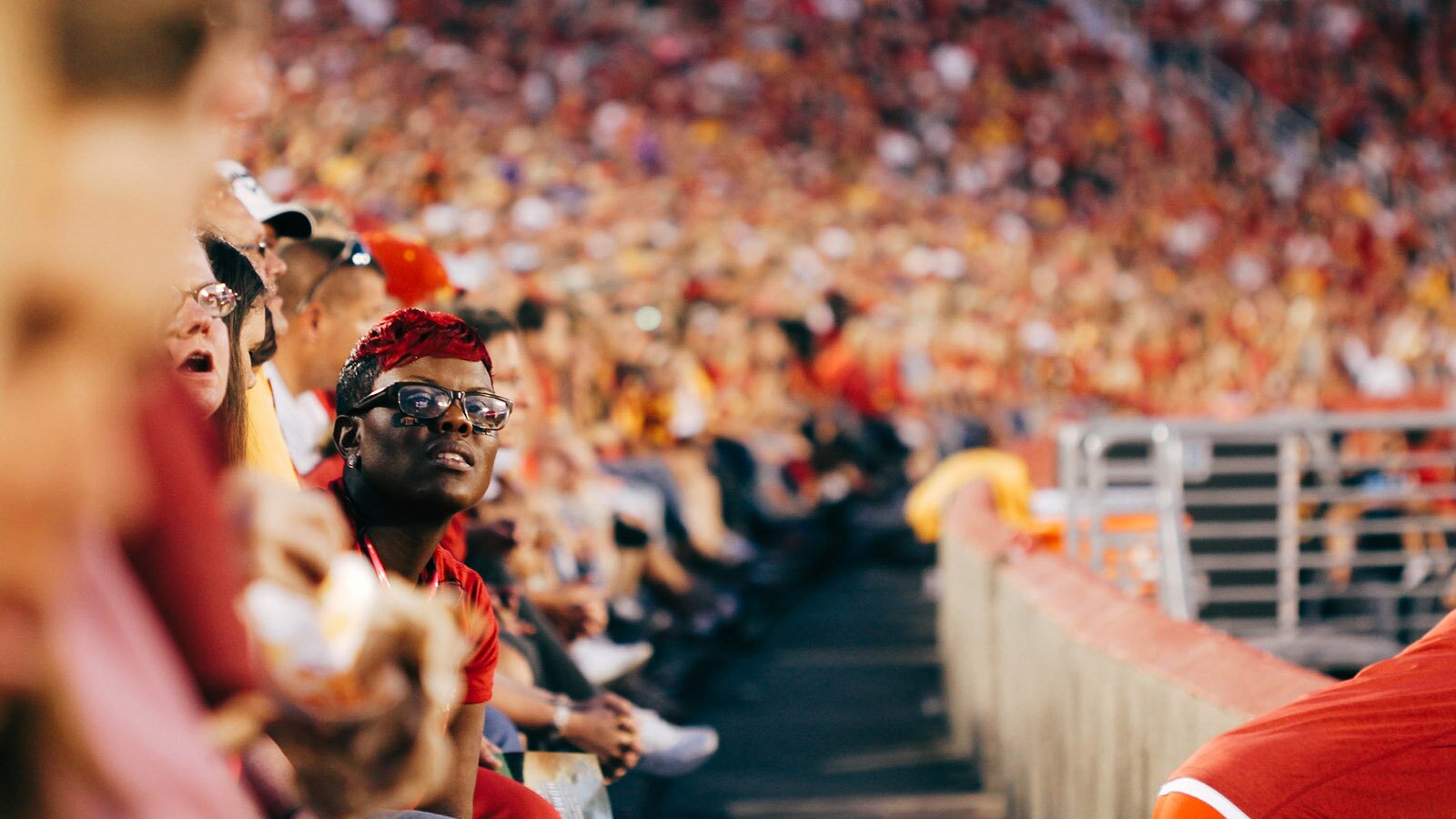 Zdjęcie przedstawia całkowicie wypełnione trybuny na wydarzeniu sportowym. Na pierwszym planie widoczna jest siedząca kobieta z czerwonymi włosami, wychylająca się do przodu.
