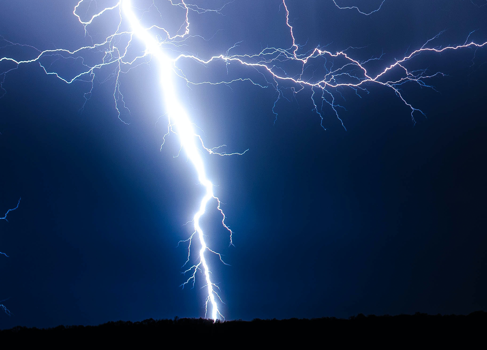 Zdjęcie przedstawia granatowe niebo rozświetlone białą błyskawicą schodzącą linią prostą w dół, a w górnej części rozgałęziającą się na boki. 