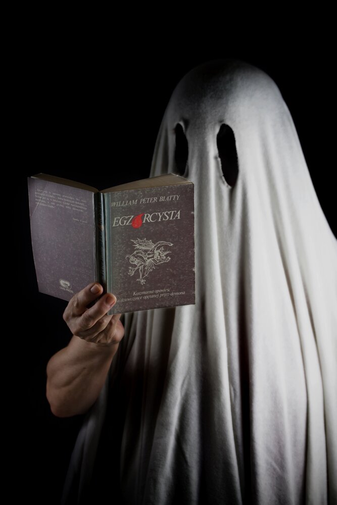 Fotografia przedstawia człowieka ubranego w prześcieradło z wyciętymi otworami na oczy. W ręku trzyma książkę pt. Egzorcysta. 