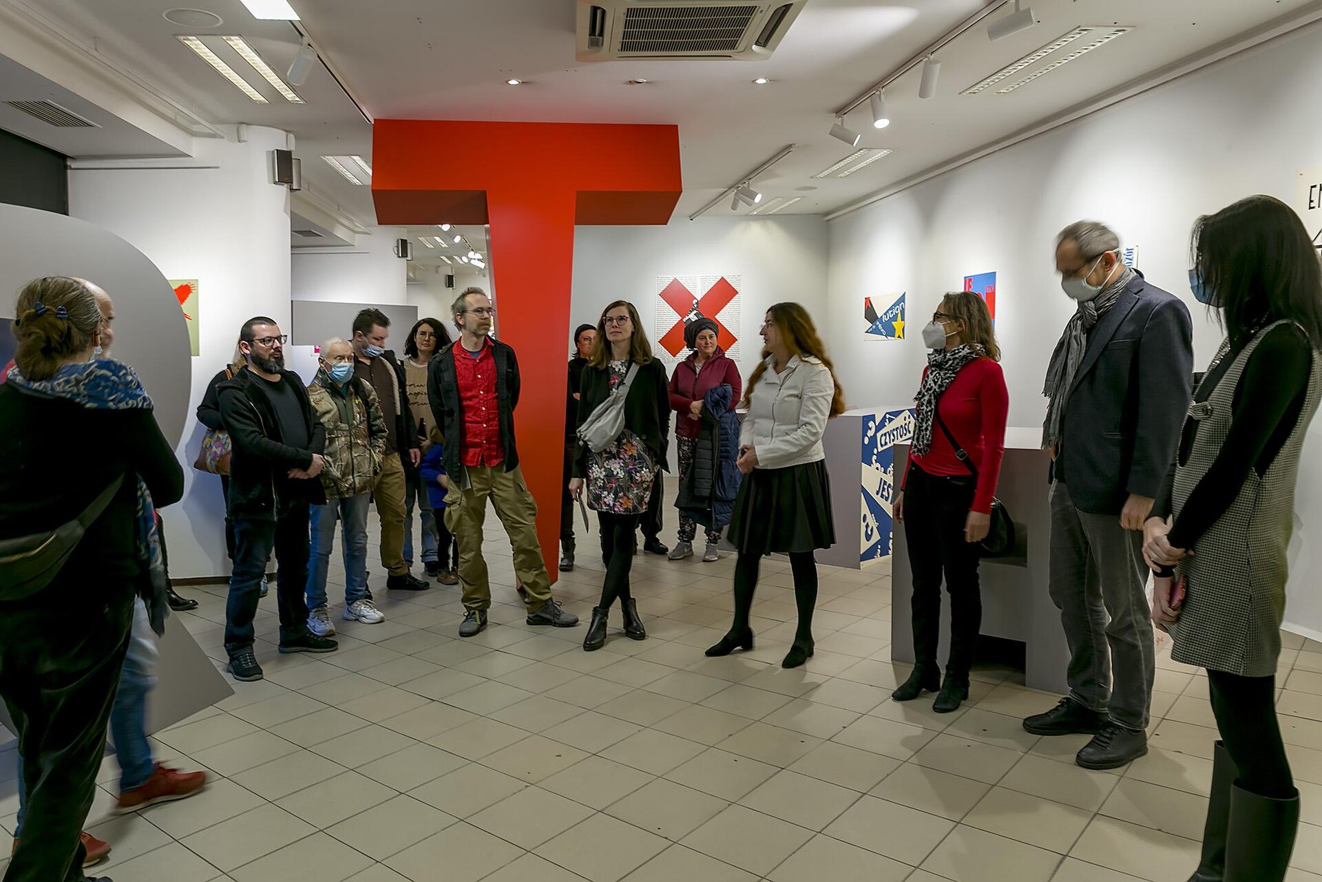 Na zdjęciu znajduje się pomieszczenie o szarej podłodze, białych ścianach i białym suficie. Na środku stoi wielka sięgająca od podłogi do sufitu figura w kształcie dużej litery "T". Dookoła litery stoi kilkanaście osób z kolorowych ubraniach. Kilka z nich ma na twarzy maseczki. Na ścianach wiszą kolorowe  prace. Na suficie są małe, białe lampki. 