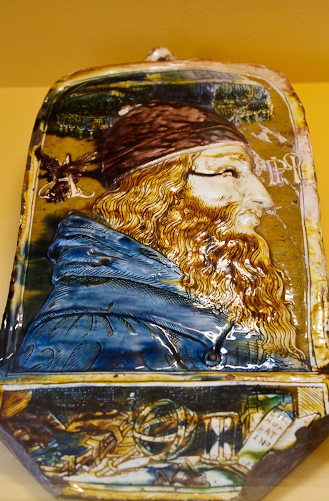 Ilustracja przedstawia ceramiczny wisior z portretem starszego mężczyzny. Ukazany jest prawy profil mężczyzny z długimi włosami, nakryciem głowy oraz gęstą brodą. Mężczyzna ubrany jest w niebieską szatę.