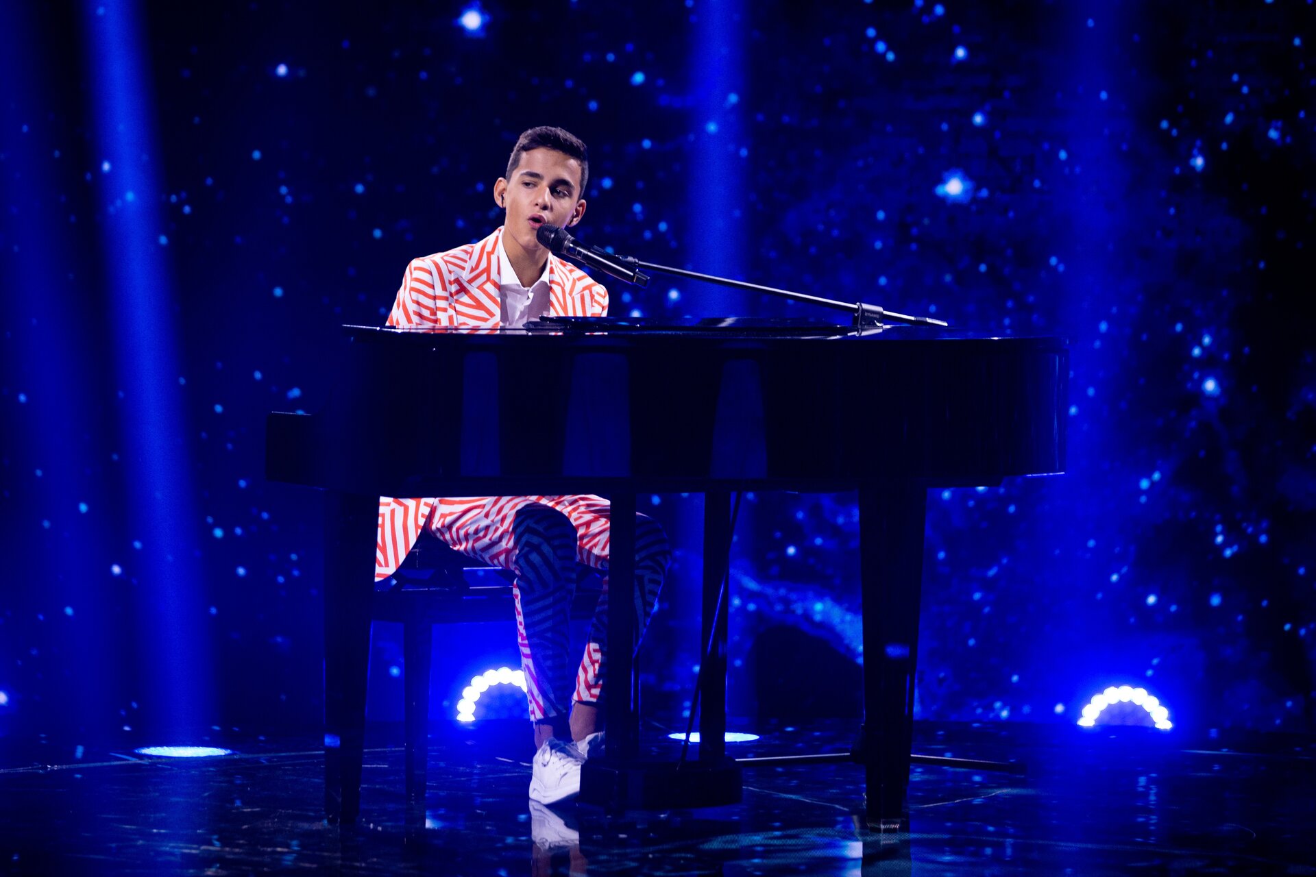 Fotografia przedstawia młodego mężczyznę, o krótkich włosach, ubranego w biało‑pomarańczowy garnitur, wykonującego utwór na fortepianie stojącym na scenie. Obok śpiewającego muzyka widoczne są słupy światła wychodzące z podłogi sceny.