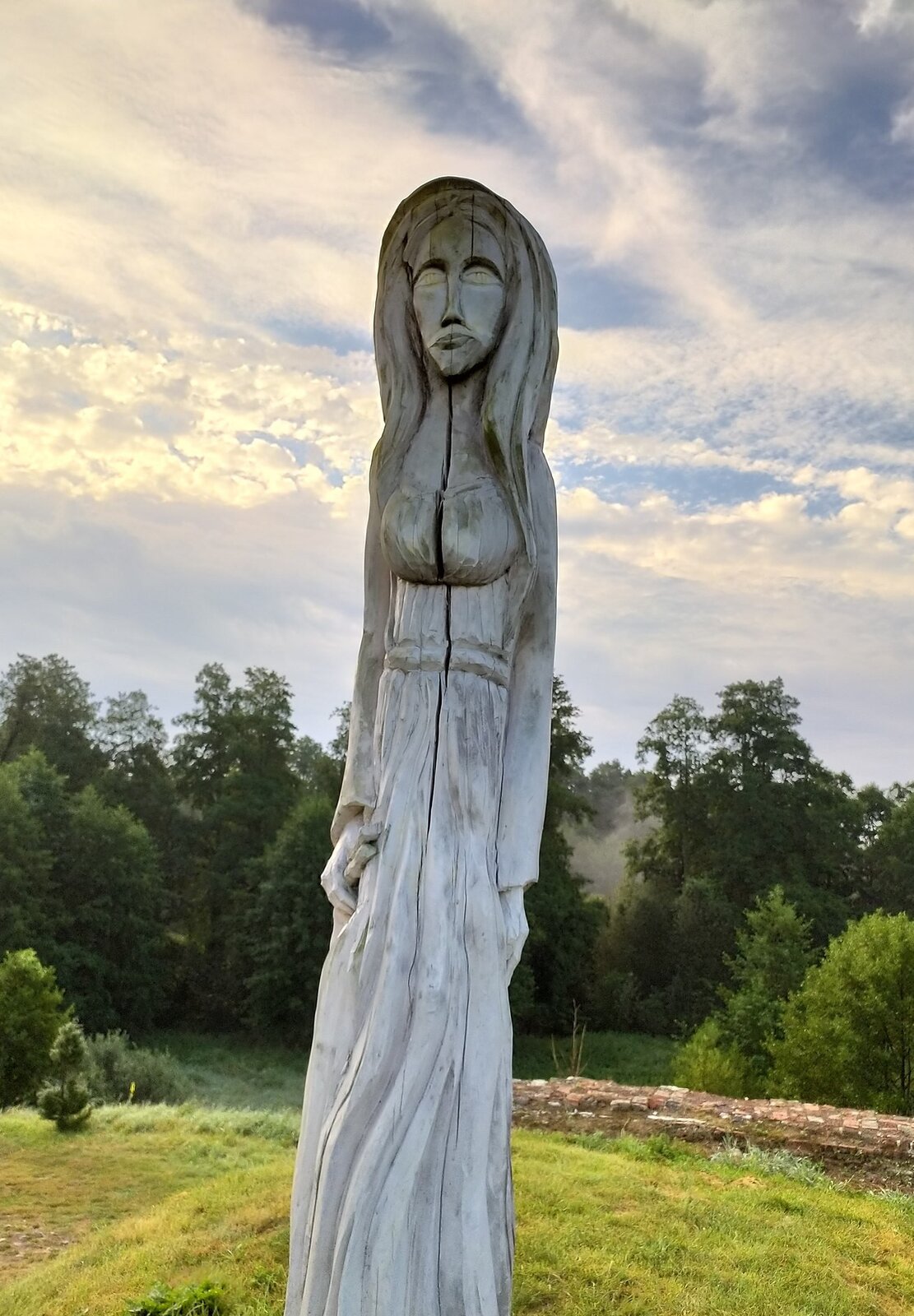 Kliknij, aby powiększyć Na kolorowej fotografii widać jasną drewnianą rzeźbę, wyobrażenie słowiańskiej bogini Mokosz. Jej postać jest bardzo wysoka i szczupła, ma duże oczy, długie włosy i obfity biust. Jej wyraz twarzy jest smutny. Rzeźba stoi na niewielkim wzgórzu, w tle las liściasty i niebo pokryte chmurami.