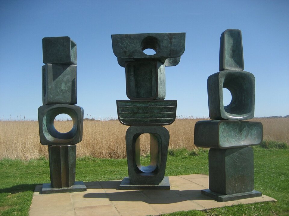 Ilustracja przedstawia abstrakcyjną rzeźbę z brązu wykonaną przez Barbarę Hepworth „Rodzina Człowiecza”. Są to trzy posągi. Każdy z nich składa się z czterech części, zbliżonych kształtem do prostopadłościanów. Każda rzeźba posiada otwór w co najmniej jednym elemencie. Bryła z otworem w każdej z rzeźb znajduje się na innym poziomie. Niektóre elementy zawierają wklęsłości. Zespół rzeźbiarski ustawiony jest na tle krajobrazu.