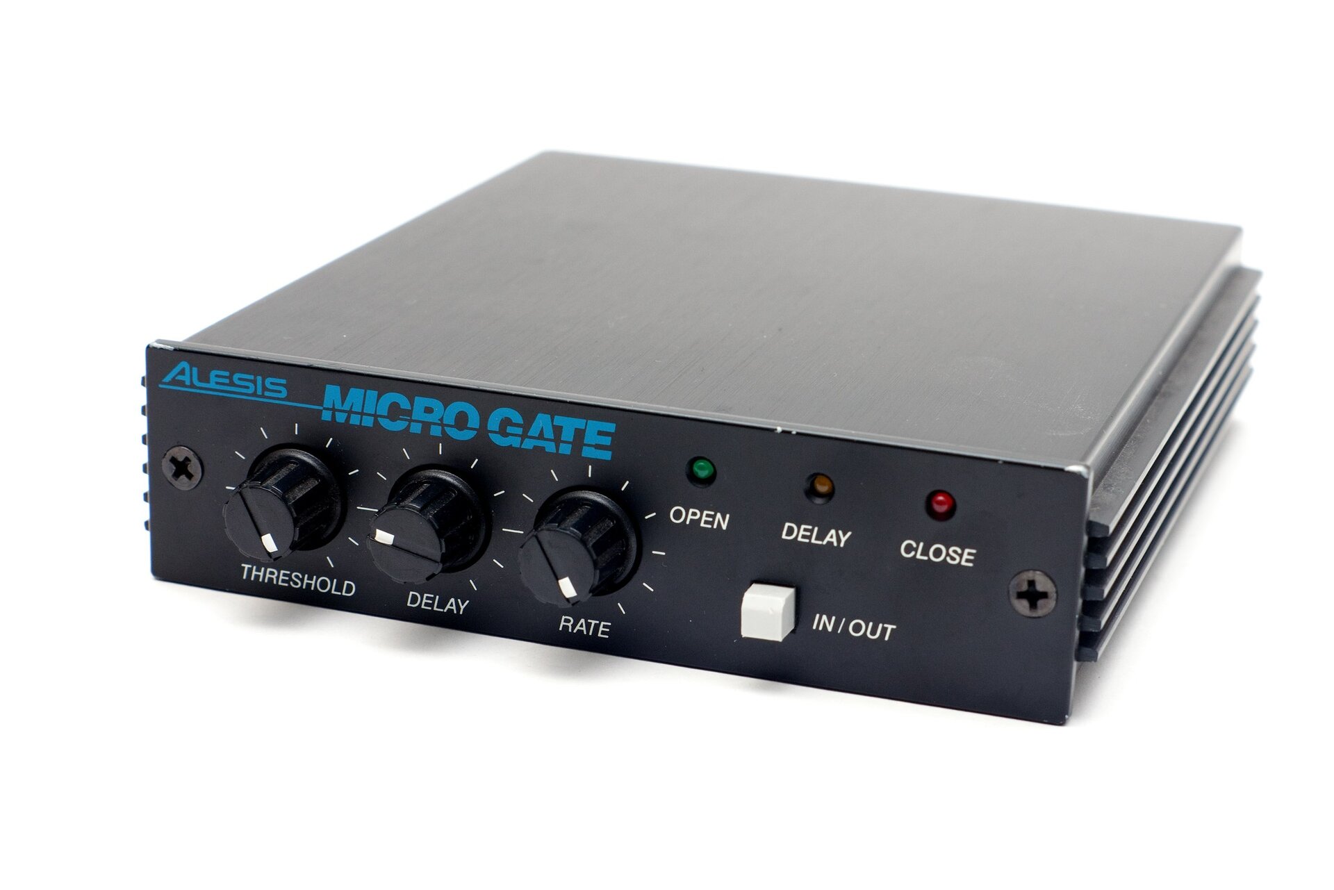 Zdjęcie przedstawia urządzenie stosowane do pracy z torem audio, w ujęciu trzy czwarte. Widać czarny prostopadłościan, na przednim boku widoczny jest panel z lampkami i pokrętłami oraz w górnej części z napisem w niebieskim kolorze Alesis Micro Gate. Poniżej napisu, od lewej trzy czarne okrągłe pokrętła threshold, delay i rate, ustawione w różnych pozycjach, a na każdym z nich mały biały prostokąt, który wskazuje ustawienie pozycji. Dookoła pokręteł krótkie białe kreski ułożone promieniście, w równych odstępach. Na prawo, w górnym polu panelu trzy kółka – lampki sygnalizacyjne, od lewej, zielone – open, czarne – delay, czerwone – close. Poniżej biały sześcian z napisem in/out.