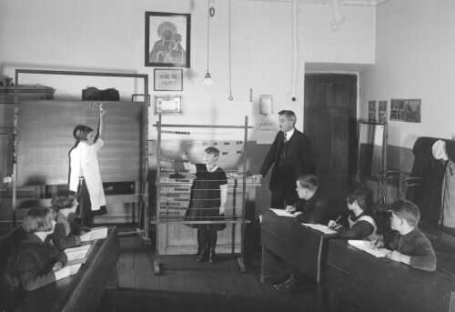 Fotografia przedstawia lekcje w jednej ze szkół. Widać uczniów siedzących w ławkach. Jedna uczennica pisze na tablicy, druga stoi przy liczydle. Wszystkiemu przygląda się nauczyciel. Nad tablicą wisi obraz Matki Boskiej. Klasa jest mała i ma kilkoro uczniów.