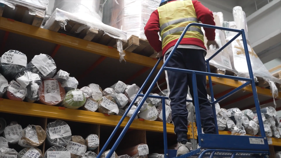 Zdjęcie przedstawia pracownika w kamizelce odblaskowej, który stoi na podeście. Podest to duże, niebieskie urządzenie. To urządzenie można przenosić po magazynie. Składa się ze schodów, barierki. Na górze jest miejsce, na którym stoi pracownik. Koło tego urządzenia jest regał z towarem.