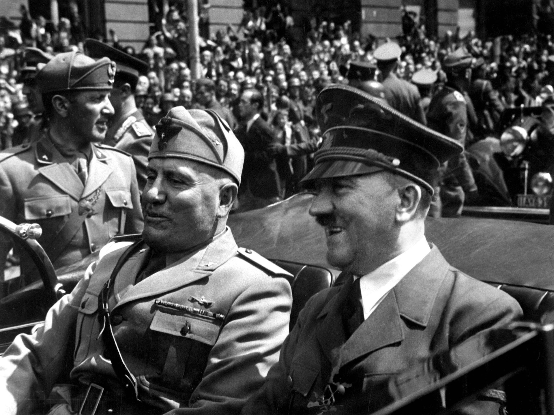 Zdjęcie przedstawia dwóch mężczyzn jadących samochodem kabrioletem. Jeden z nich to niemiecki żołnierz, ma wąs pod nosem, to Adolf Hitler. Drugi z nich, to włoski żołnierz, Benito Mussolini. W tle widać tłumy żołnierzy. 