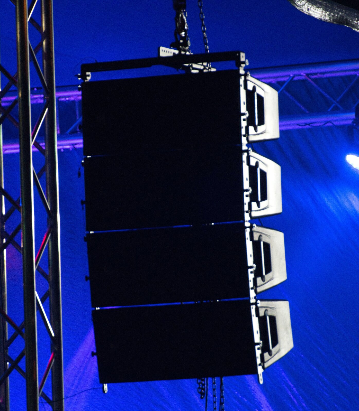 Zdjęcie przedstawia klaster nagłośnieniowy, czyli zestaw głośników. Widoczny jest duży, czarny i zawieszony na czarnym łańcuchu, prostokąt, z tyłu którego znajdują się cztery czarne trapezy, z ażurowymi elementami w dolnej części. Po lewej stronie widoczne jest rusztowanie w formie dwóch pionowych czarnych rurek, pomiędzy którymi zygzakowato biegną cieńsze czarne rurki. W tle widoczne jest rusztowanie w formie poziomych rurek, podświetlonych na fioletowo, pomiędzy którymi zygzakowato biegną cieńsze rurki. Po prawej stronie u góry widoczny ucięte białe koło – lampa. Wszystkie elementy widoczne są na fioletowym tle.