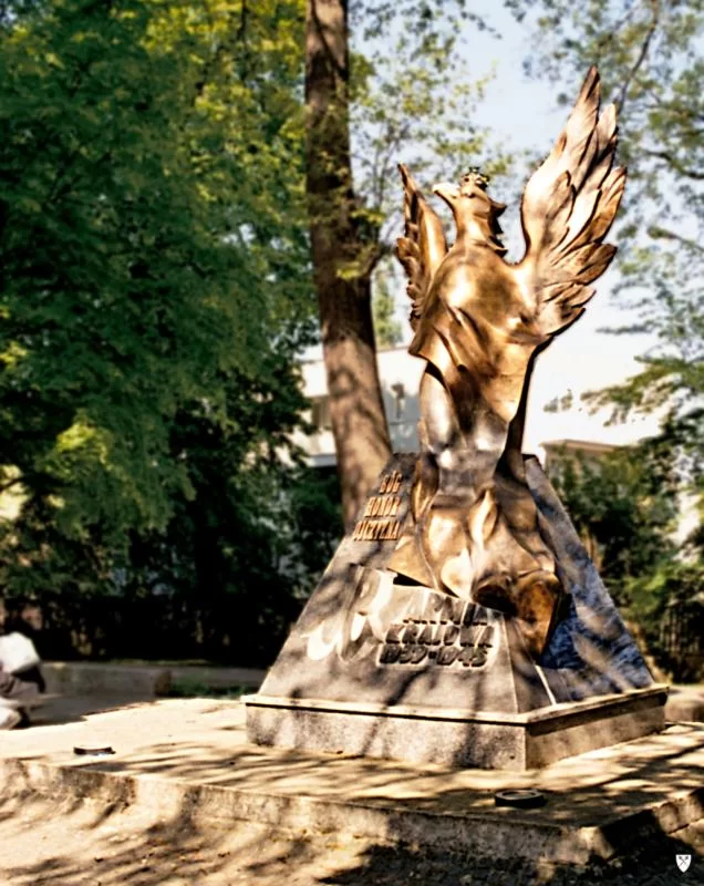 Kliknij, aby powiększyć Zdjęcie. Na podeście znajduje się niewielki monument w postaci trapezu ze spoczywającym na nim orłem. Orzeł parzy w lewo, nosi koronę i rozpościera skrzydła. Na pomniku wygrawerowano napis: Bóg, Honor, Ojczyzna i Armia Krajowa 1939 - 1945. W tle drzewa.