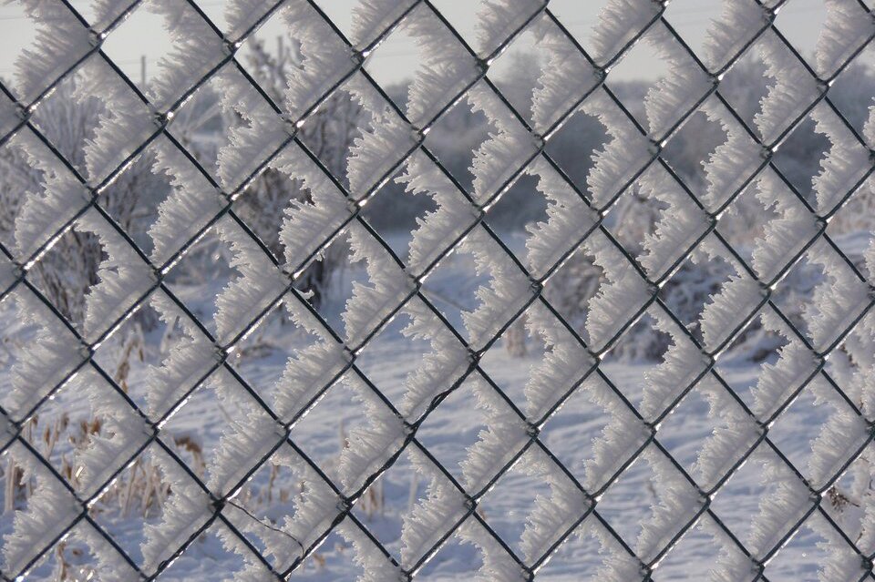 Fotografia barwna przedstawiająca zimowy, mroźny dzień. Na ilustracji szadź, która pokrywa całą powierzchnię siatki, z której zbudowane jest ogrodzenie.