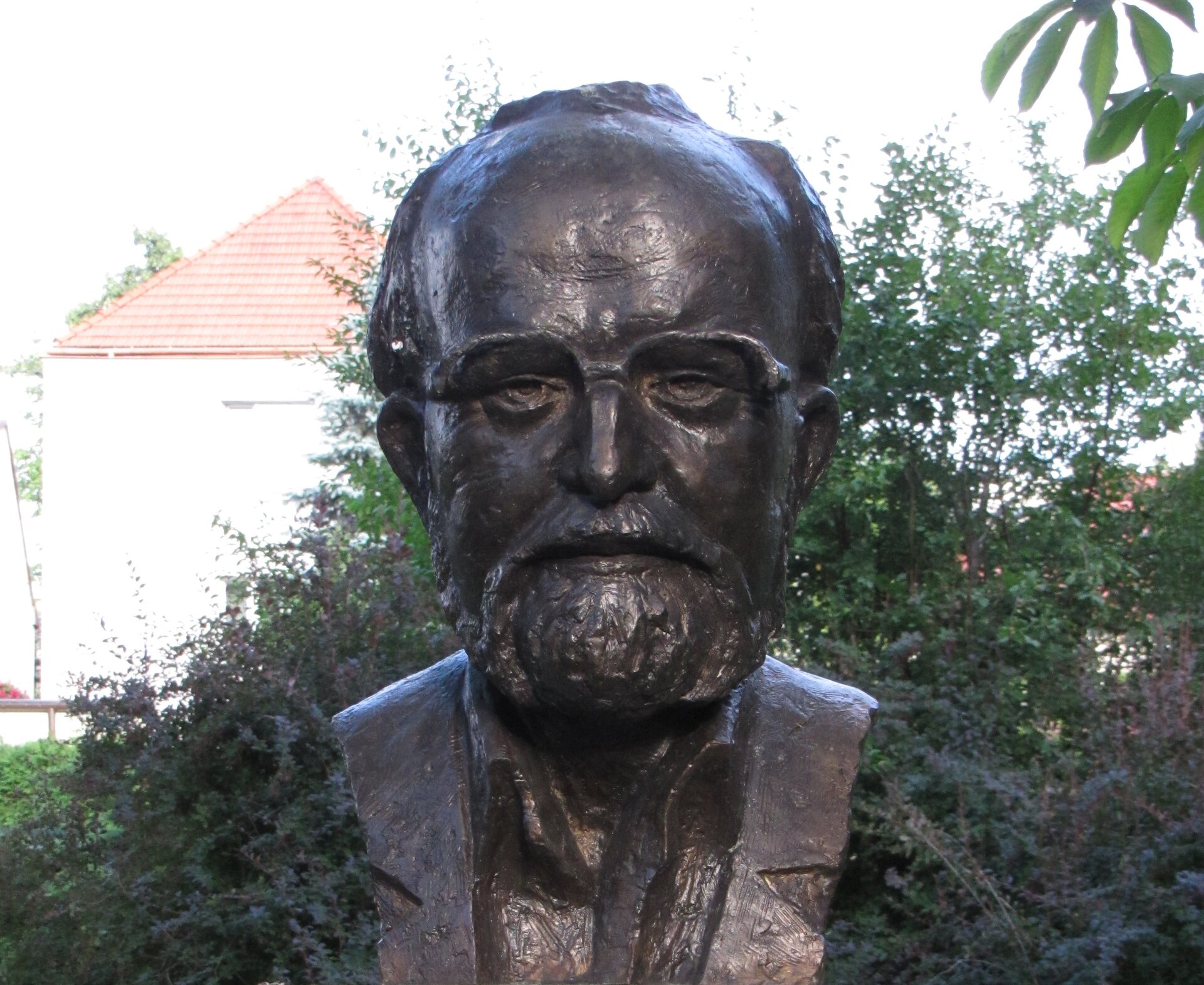 Fotografia przedstawia popiersie Krzysztofa Pendereckiego wykonane przez Leszka Kurkowskiego. Popiersie jest portretem starszego mężczyzny o łysiejącej głowie, z włosami po bokach i na czubku głowy. Ma on szeroko otwarte oczy i ze spokojem spogląda na widza. Ma brodę oraz wąsy. Jego skóra jest pełna pomarszczona oraz niedoskonałości. Popiersie stoi w parku, na tle krzewów oraz budynku.  