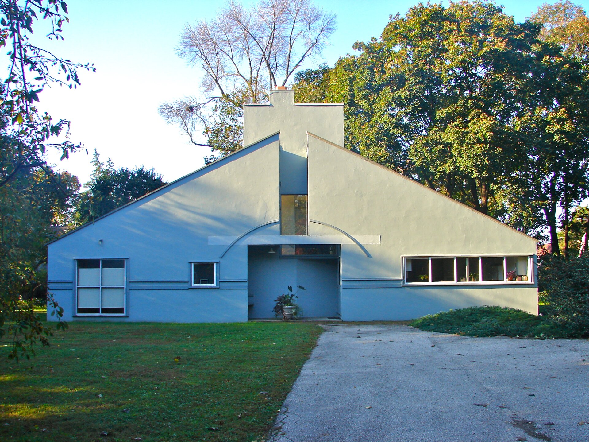 Fotografia przedstawia szary dom z jednospadowym dachem z szerokim kominem. Po środku domu, tuż pod kominem znajduje się wycięcie z oknem, a niżej znajduje się wejście główne budynku. Po prawej stronie ukazane jest długie okno. Po lewej stronie przedstawiono dwa okna – mniejsze oraz większe. Dom otoczony jest drzewami oraz krzewami.