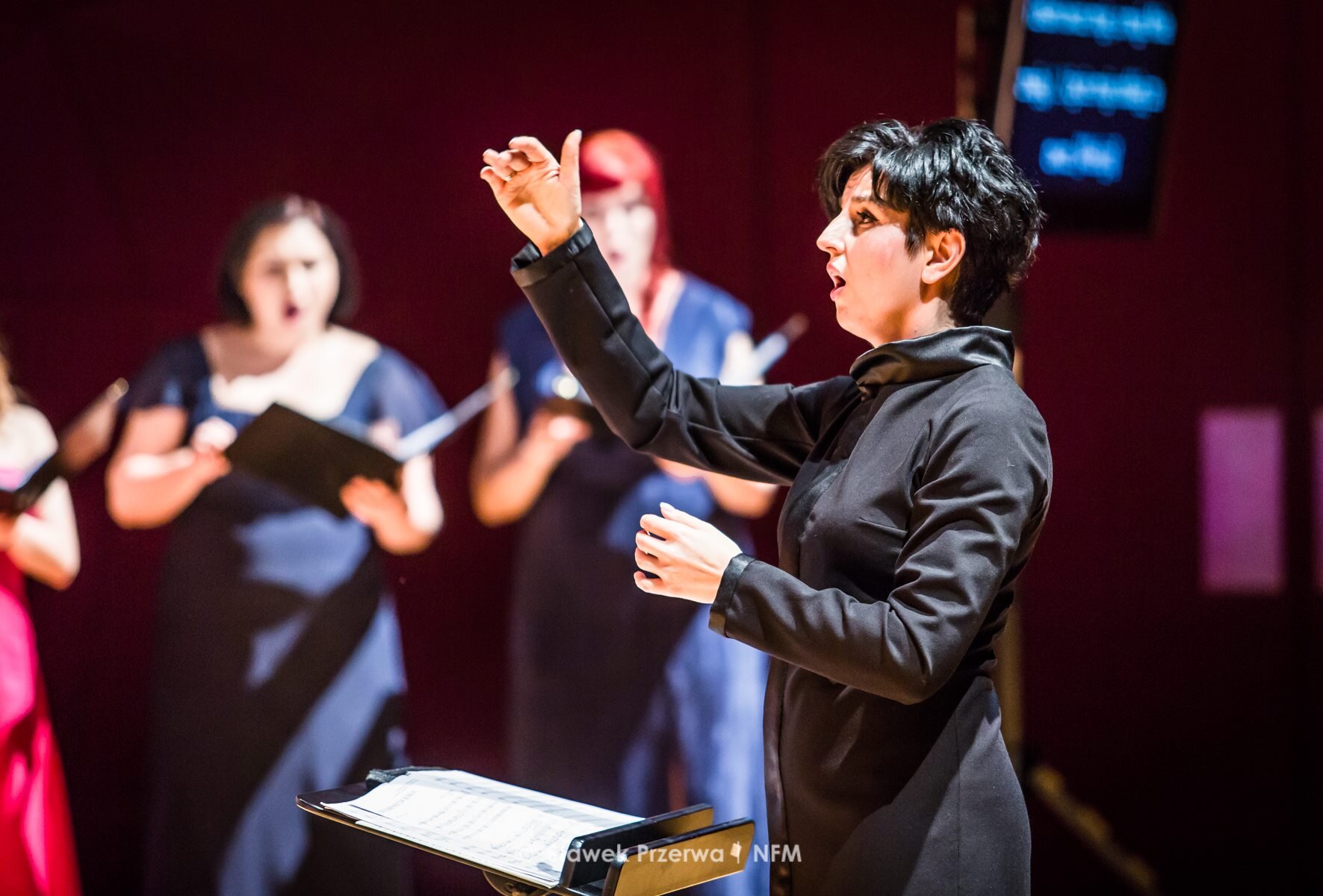Fotografia Agnieszki Franków‑Żelazny – dyrygent chóralny i orkiestrowy Narodowego Forum Muzyki we Wrocławiu Fotografia przedstawia kobietę‑dyrygent, panią Agnieszkę Franków‑Żelazny w trakcie koncertu. Kobieta została przedstawiona z profilu, ma krótkie ciemne włosy, ubrana jest w czarny płaszcz. Jej prawa ręka jest uniesiona do linii głowy, a lewa – umieszczona przed klatką piersiową. Przed kobietą ustawiony został stolik z partyturą utworu. W tle stoją kobiety - chórzystki.