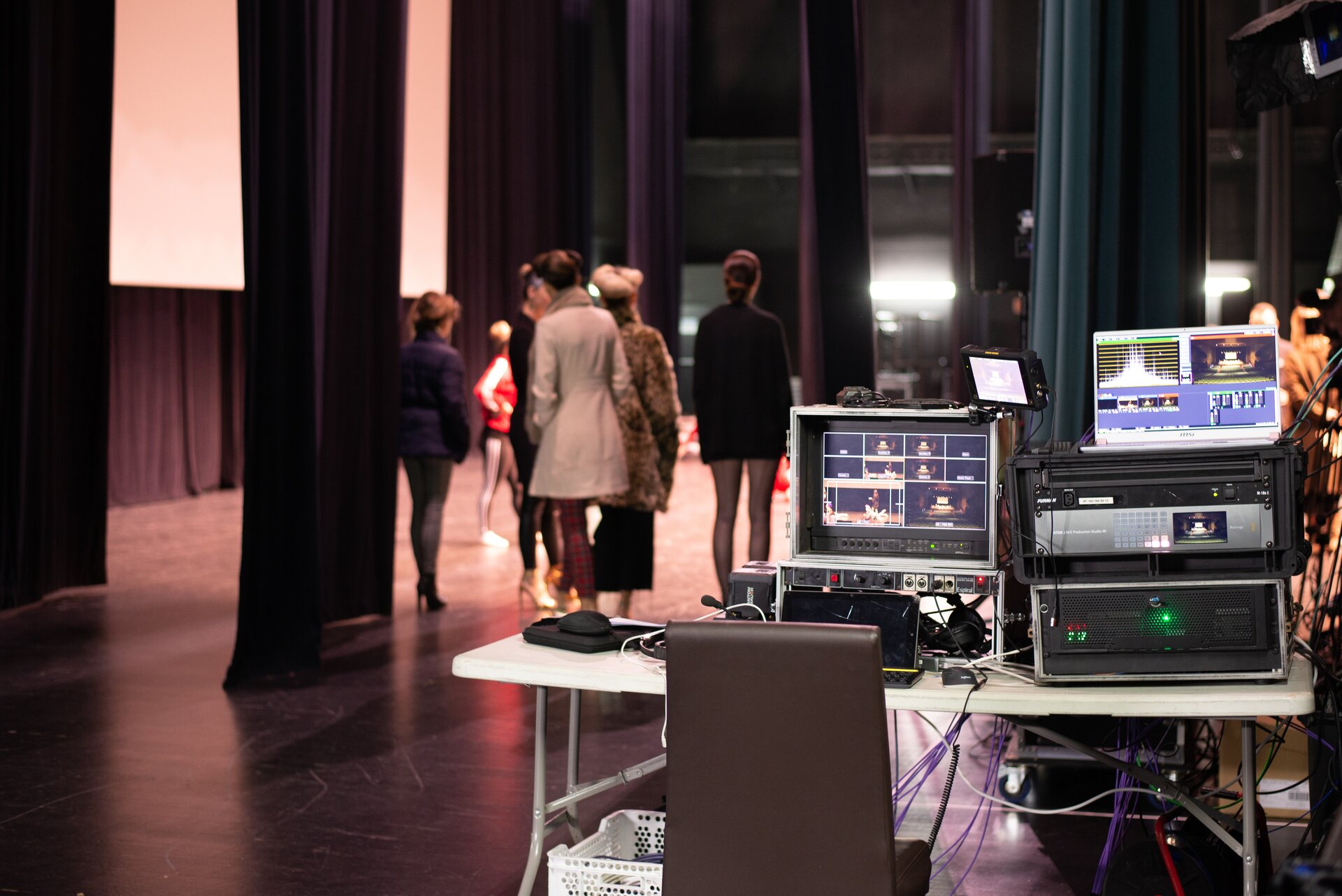 Zdjęcie przedstawia stolik, na którym ułożony jest sprzęt elektroniczny. Na ekranach wyświetla się widok na scenę i widownię. Na laptopie wyświetla się ścieżka dźwiękowa. Na sprzęcie świecą się diody, podłączone są kable. W tle znajdują się kulisy, w których stoją ludzie, a za nimi jest scena.