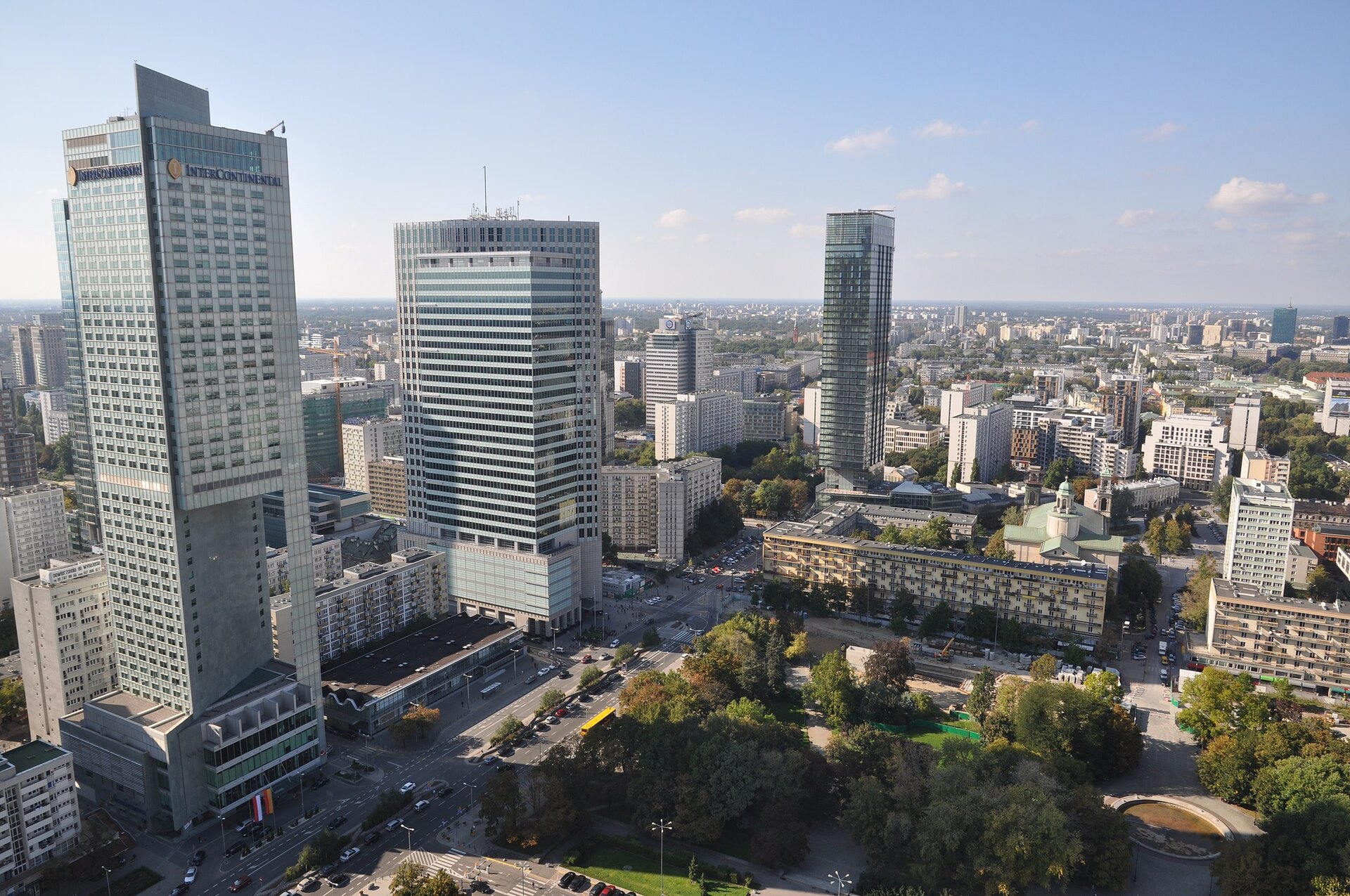 Zdjęcie przedstawia panoramę miasta. Na pierwszym planie znajduje się plac obsadzony drzewami. Po lewej stronie znajdują się trzy wysokie wieżowce, które górują nad pozostałymi budynkami. Po prawej stronie rozciąga się widok na kamienice i blokowiska.
