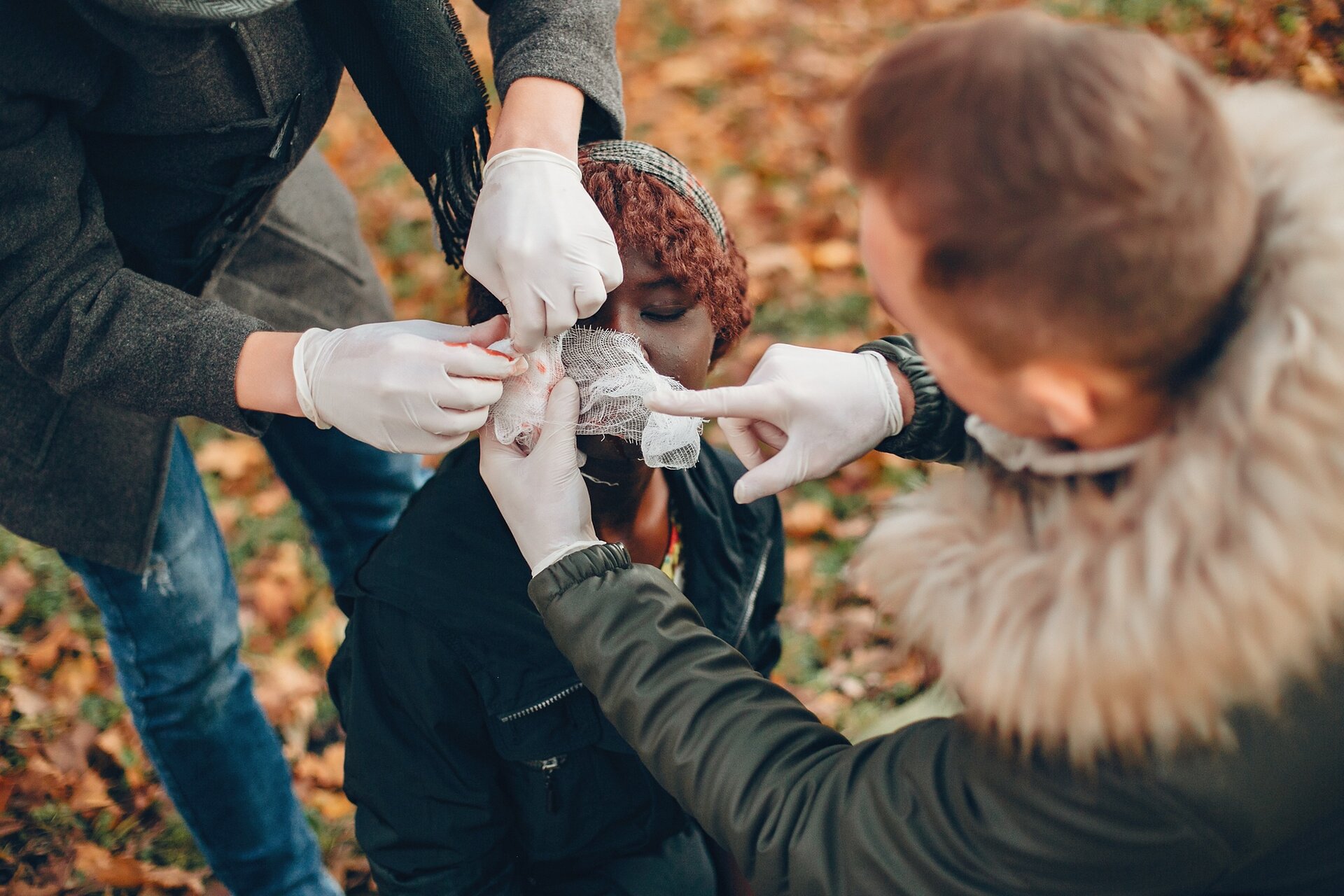 Ilustracja przestawia dziewczynę z raną twarzy, która jest zaopatrywana za pomocą środków opatrunkowych przez dwie osoby w rękawiczkach jednorazowych.