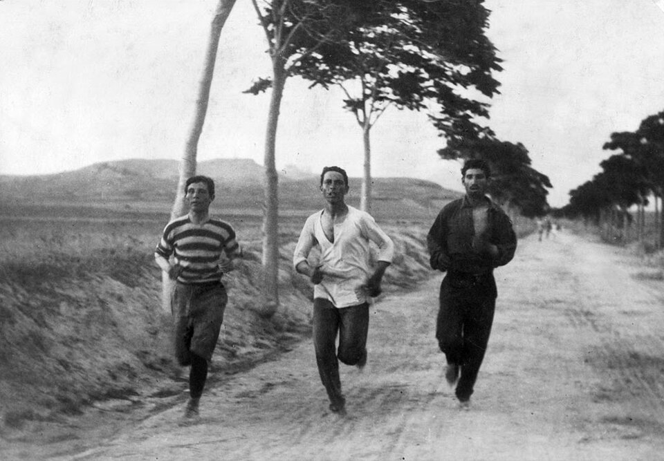 Fotografia autorstwa Burtona Holmesa pod tytułem „Trzej atleci przygotowujący się do maratonu w czasie igrzysk w Atenach w 1896 r”. Na fotografii uwieczniono trzech mężczyzn biegnących po nieutwardzonej drodze. Każdy ze sfotografowanych mężczyzn jest młody, ma ciemną karnację oraz ciemne włosy. Mężczyzna z lewej strony ubrany jest w dwukolorową pasiastą koszulkę z podwiniętymi rękawami oraz długie spodnie. Mężczyzna biegnący po środku ubrany jest w długą zwiewną koszulę oraz ciemne, długie spodnie. Trzeci biegacz ubrany jest w czarną koszulę, czarne spodnie, dodatkowo na dłonie założone ma czarne rękawiczki. W tle znajdują się drzewa oraz góry.
