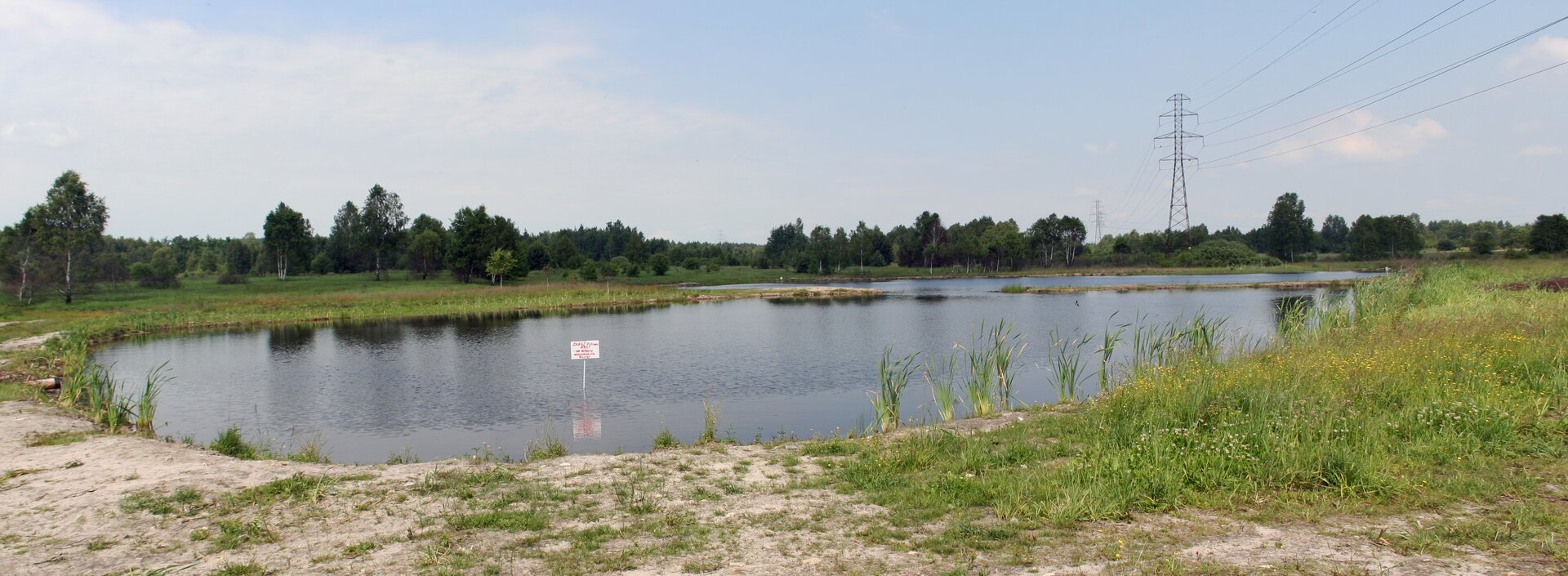 Fotografia przedstawiająca lej wypełniony wodą. Ma średnicę około kilkudziesięciu metrów, przypomina małe jezioro. Dookoła  płaski teren porośnięty trawą, w oddali drzewa.