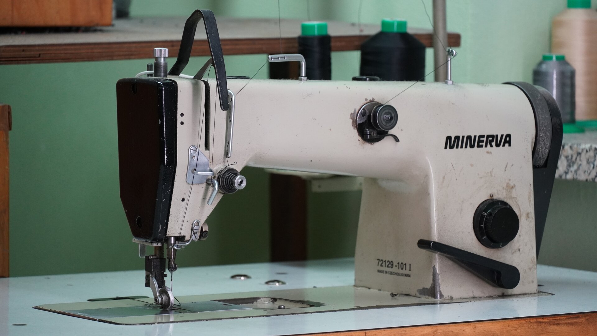 Zdjęcie przedstawia białą maszynę płaską do szycia z czarnymi elementami. Z przodu ma napis MINERVA. W górnej części założone szpulki z nićmi. Nagranie tożsame z treścią.