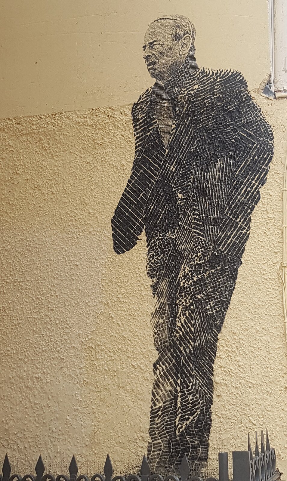 Zdjęcie przedstawia naścienny mural. Znajduje się na nim sylwetka dojrzałego mężczyzny. Ma krótkie, zaczesane na bok włosy oraz zmarszczki na twarzy. Mężczyzna mruży oczy. Ubrany jest w garnitur z krawatem. Trzyma ręce w kieszeniach spodni.