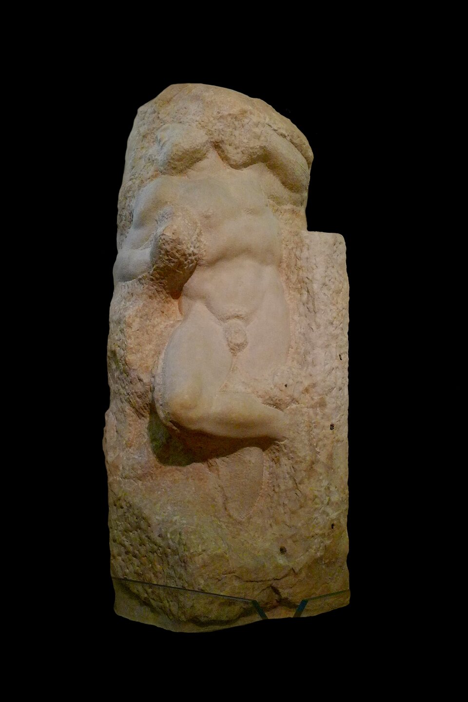 Niedokończona marmurowa rzeźba przedstawiająca nagą, muskularną postać mężczyzny wyłaniającą się z surowego bloku kamienia. Sylwetka jest skręcona, z jedną nogą zgiętą i wysuniętą do przodu, a ramiona uwięzione w nieociosanej masie marmuru. Górna część tułowia i uda są bardziej wygładzone, z wyraźnie zaznaczonymi mięśniami, podczas gdy reszta bryły pozostaje chropowata, z widocznymi śladami narzędzi. Całość ukazuje kontrast między dopracowaną anatomią a nieobrobioną strukturą kamienia na czarnym tle.