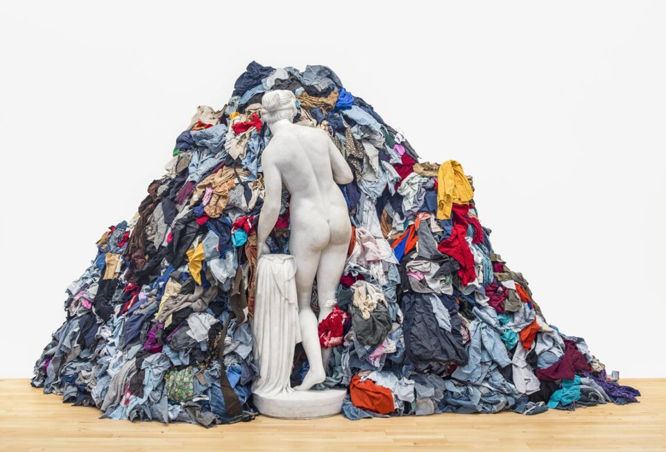 Ilustracja w formie prostokąta przedstawia posąg kobiety o krótkich, spiętych włosach, który odwrócony jest tyłem do widza. Kobieta jest naga, a w lewej ręce trzyma szatę. Kobieta stoi na lekko ugiętych nogach, a jej ciało pochylone jest delikatnie w lewą stronę. Kobieta wchodzi w stertę rozrzuconych ubrań. Są one w wielu kolorach. Dzieło ustawione jest w pomieszczeniu o białych ścianach i jasnej, drewnianej podłodze. 