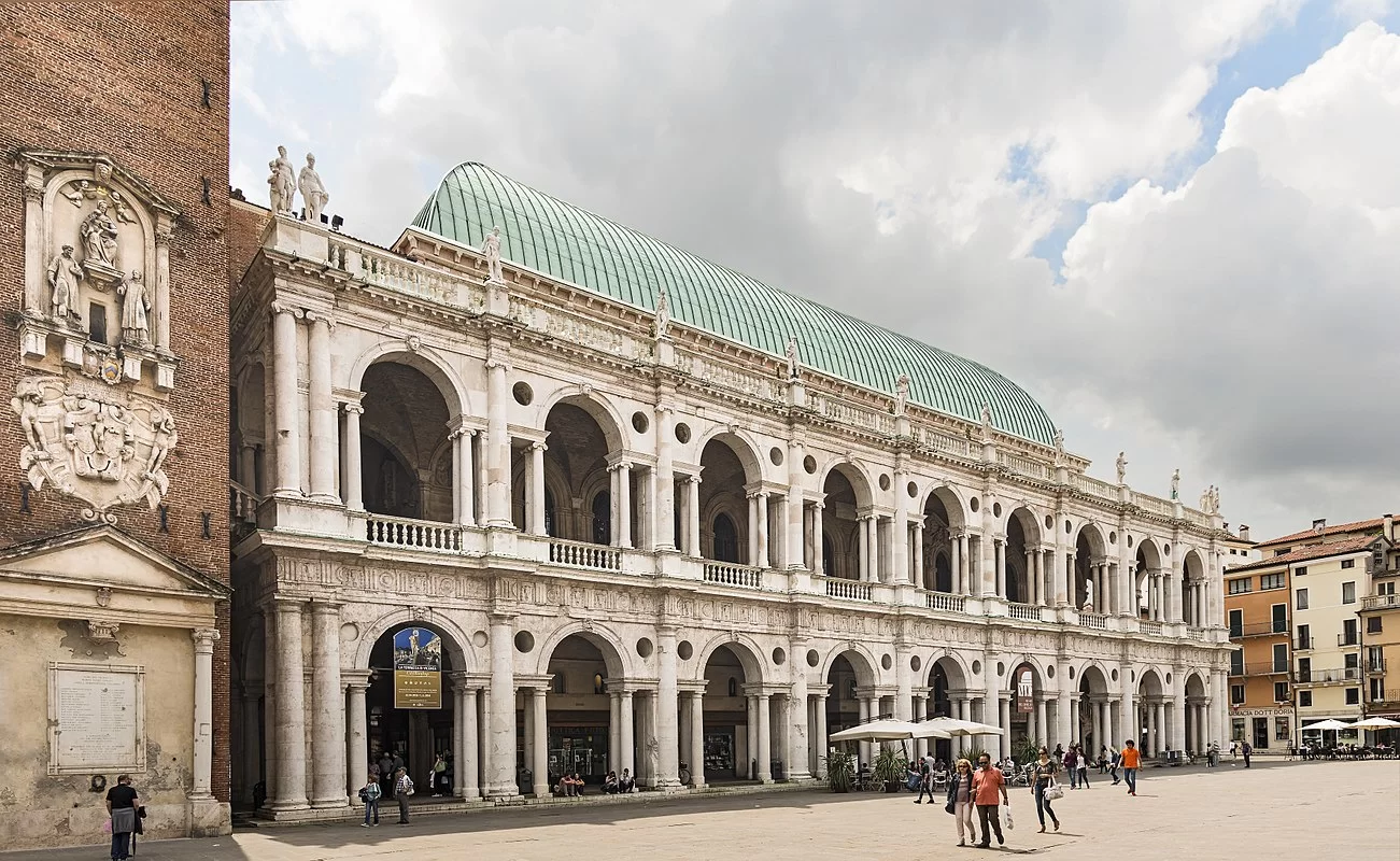 Zdjęcie przedstawia fragment renesansowej bazyliki na centralnym placu Vicenzy. Budynek ma dwie kondygnacje, na każdej znajduje się dziewięć arkad. Arkady oddzielają do siebie kolumny. Całość zwieńczona jest półokrągłym dachem.
