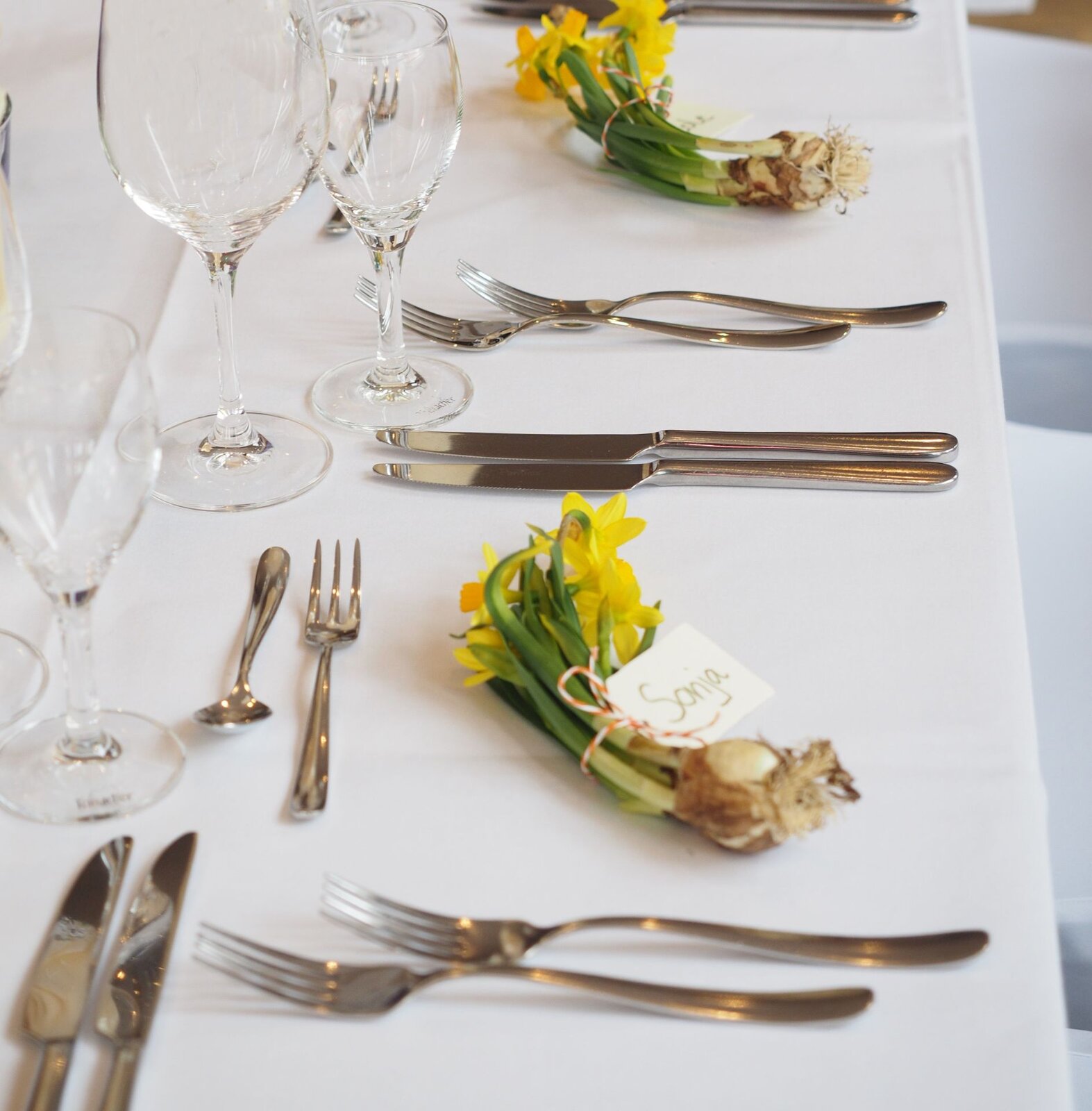 Zdjęcie przedstawia fragment zastawionego stołu na sali bankietowej. Każdy gość ma po swojej prawej dwa widelce, po lewej dwa noże, nad talerzem widelec deserowy i łyżeczkę. W miejscu talerza znajdują się bukiety żółtych żonkili.  W większym kieliszku jest kremowa serwetka zwinięta w wachlarzyk. 
