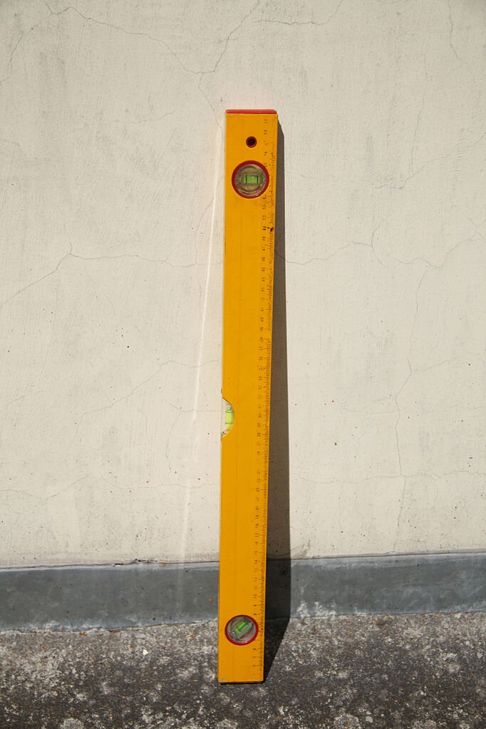 Zdjęcie przedstawia poziomicę. Jest to podłużne narzędzie z trzema okienkami, w których znajduje się libelle rurkowe, pozwalające na wyznaczenie poziomości lub pionowości płaszczyzn.