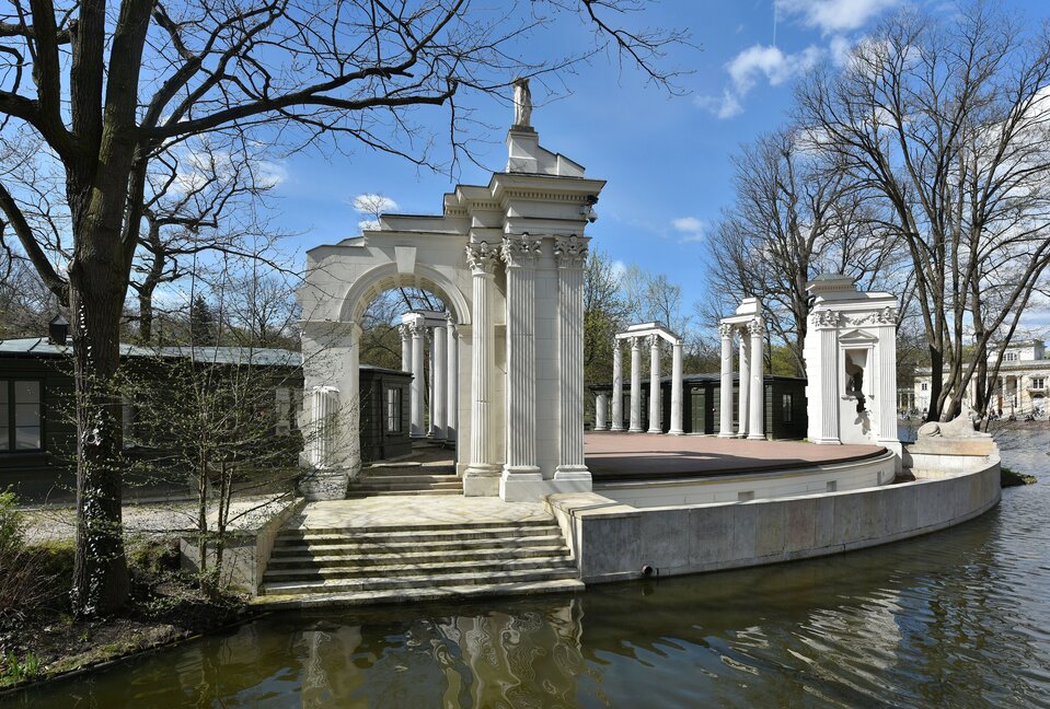Ilustracja przedstawia amfiteatr w łazienkach w Warszawie, wzorowany na teatrze w Herkulanum. Półkolista widownia oparta jest na arkadach. Wieńczy ją rząd kamiennych rzeźb przedstawiających szesnastu dramaturgów. Scena, położona na sztucznej wyspie otoczona jest sztucznymi ruinami, wzorowanymi na architekturze Forum Romanum. Po lewej stronie za sceną ukryty znajduje się parterowy budynek.
