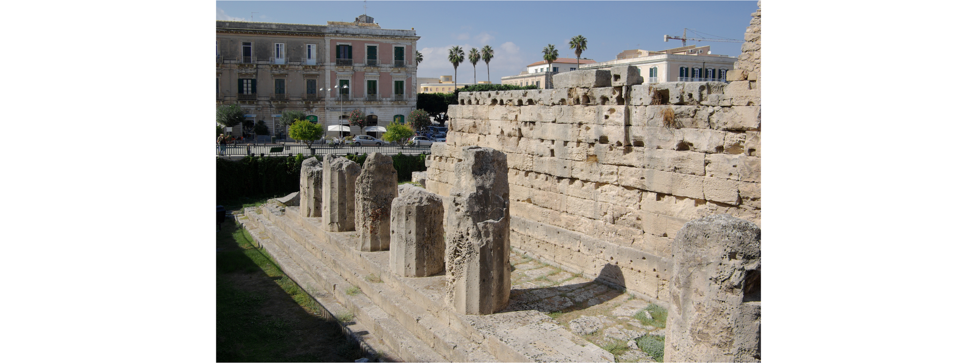 Zdjęcie przedstawia fragment starożytnych ruin miasta Syrakuzy. W tle widoczny jest kilkupiętrowy budynek.