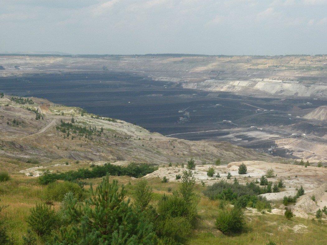 Ilustracja przedstawia ogromy obszar czarnej ziemi, który znajduje się pomiędzy wzniesieniami kopalni. Na pierwszym planie widoczna jest roślinność: trawy i krzewy pokrywające zbocza wzniesień. Wzniesienia mają kolor jasno-brązowy. W tle widoczne jest niebo pokryte chmurami. 