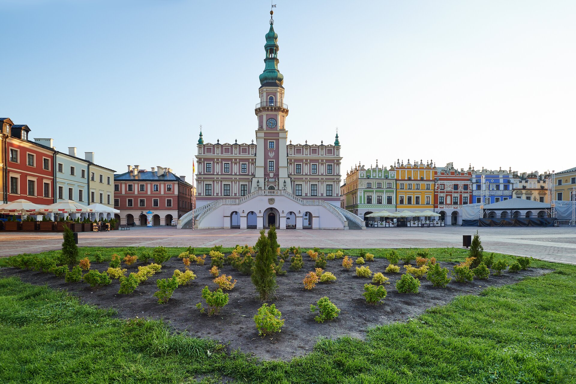 Ilustracja przedstawia zdjęcie Zamościa. Ukazuje widok na ratusz i kamienice. Jest to ujęcie z perspektywy centralnej. Przed ratuszem znajduje się plac rynku z wytyczonym zieleńcem. Ratusz ustawiony jest w centrum widoku. Prowadzą do niego podwójne schody. Frontową, trzykondygnacyjną ścianę przecina masywna wieża zegarowa, zwieńczona wysokim hełmem. Ścianę wieńczy dekoracyjna attyka. W narożach znajdują się małe wieżyczki, również kryte hełmami. Po obu stronach ratusza znajdują się kamienice, których elewacje są kolorowe. Przed budynkami ustawione są ogródki restauracyjne z parasolami.